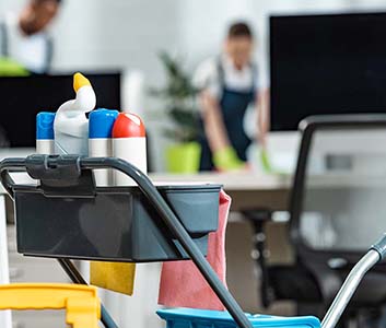 a janitor in the background cleaning a work desk along with cleaning tools in trolley